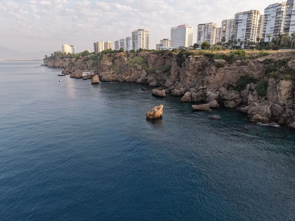Antalya: Scenic view of Antalya's coastline with rocky cliffs and urban skyline under a cloudy sky.